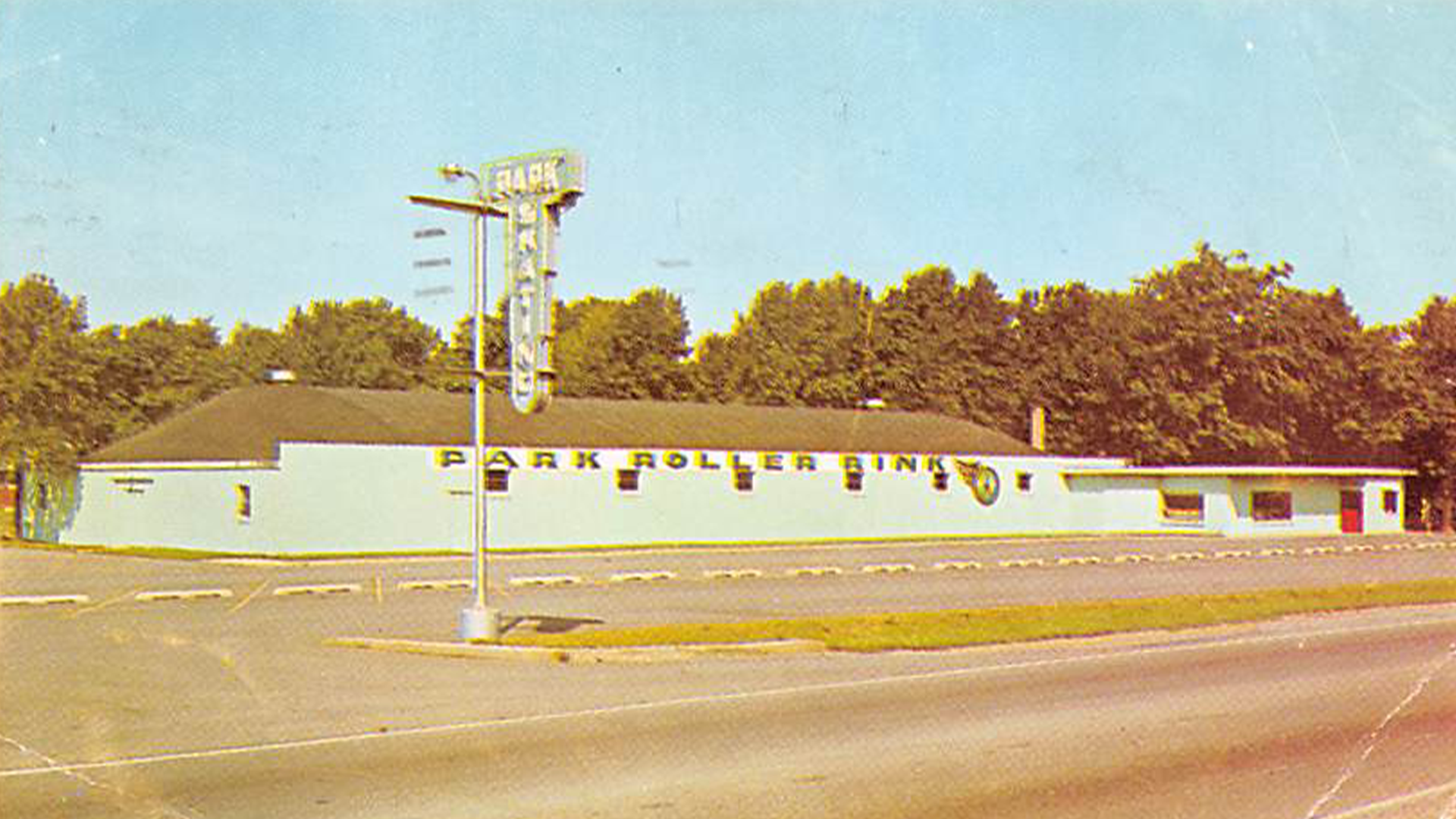 An image from the 1970's of the exterior of a roller rink, like those featured in the film ' All Skate'. The picture was taken on a bright sunny day; the parking lot of the roller rink is completely empty and a sign out front that says ' Park Skating'.