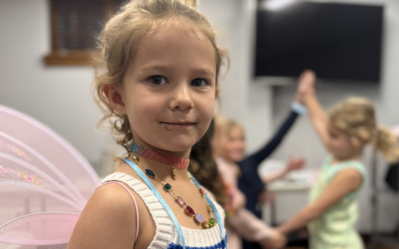 A young girl wearing pink fairy wings and a beaded necklace is smiling into the camera while other girls in the Jr. sewing class play in the background