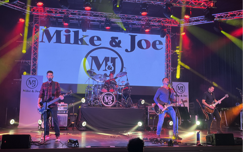 Members of the band 'Mike & Joe' performing on stage. The band name and logo are projected onto a large screen behind the band and there are red and yellow spotlights shining down on the stage.