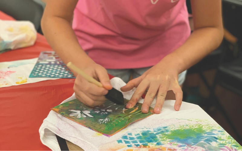 A camper of the Design & Craft camp with Vee Gunn is using a stencil to paint a pattern on a t shirt.