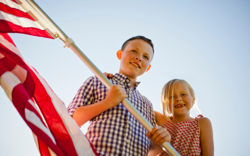 a boy holds a large American flag with a younger blonde girl standing beside him