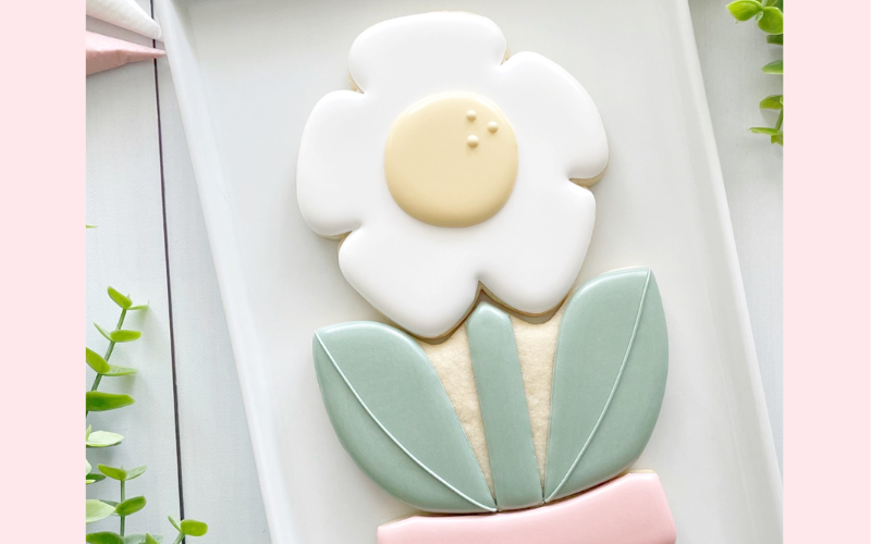 A sample cookie from the Mom and Me cookie decorating class. The cookie is in the shape of flower. It has white petals, sage green leaves and is coming out of a pink pot. The cookie is laying on a white linen napkin.