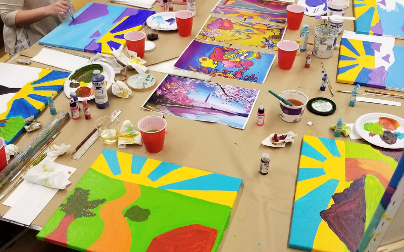 An image from a Palette and Pours class at Gorton. A long table is covered with paper; there are printed images int he middle and along the sides are canvases that are works in progress. Each painting has a bright sun in the middle surrounded by blue sky. There are also cups of water, palettes, paints and brushes all over the table.