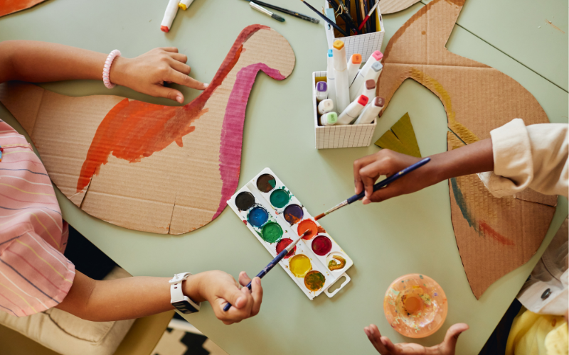 Two campers at the Creativity Hack are at a table painting pieces of cardboard that have been cut out into the shapes of animals. The camera is above the table, the campers hands are visible and there is a paint pallet on the table between them.