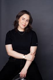 a photo of stand up comedian Kate Kincaid who is opening for Emily Catalano. Kate is dressed in all black, she has her head tilted to the right and she is smiling at the camera
