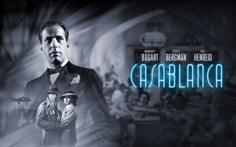 A film poster for 'Casablanca'- the image is in black and white; There is a large image of actor Humphrey Bogart in a tuxedo with the faint image of a band behind him. Below that is an image of actor Paul Henreid standing with actress Ingrid Bergman. They are clinging onto each other and looking off into the distance.