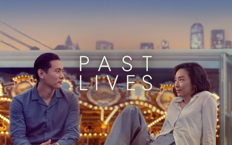 An image from the film ' Past Lives'. A man and a woman sitting in front of a carousel with a city skyline behind them. The man and woman are looking intently at each other.