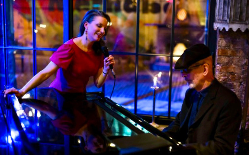 Jazz Vocalist Petra van Nuis is standing in front of a window in a dimly lit room- to her left is pianist Dennis Luxion. Petra is wearing a red dress, leaning slightly over the piano and singing into a microphone. Dennis is seated and looking down at the keys as he plays the piano.