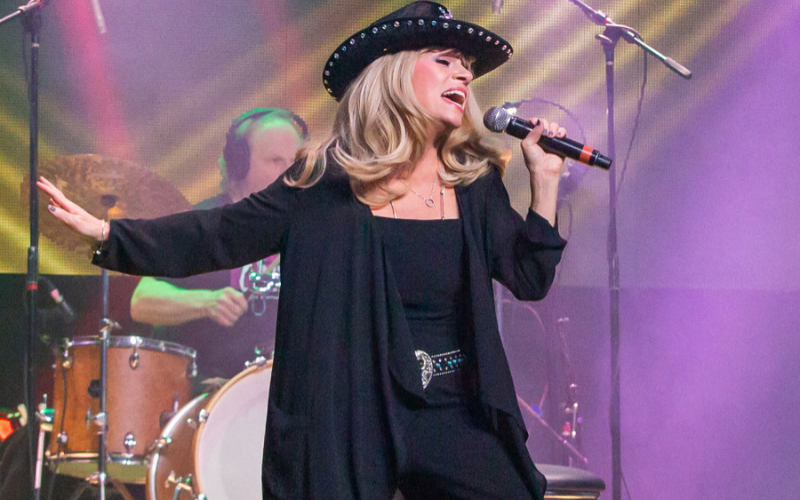 An image of Annie Aiello as performing on stage as Olivia Newton John: she has a black leather jacket she is wearing a black cowboy-style hat in the style of 'Xanadu'