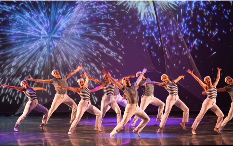 A performance image of Giordano Dance Company on stage- they are all dressed as sailors with white pants , blue striped shirts and sailor hats. Each dancer has their arms outstretched and their heads tilted back. Behind them, images of fireworks are being projected.