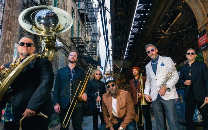 The members of the Lowdown Brass Band are posing with their instruments and looking at the camera. They are nicely dressed and in an alley with buildings and fire escapes visible in the background.