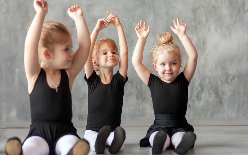 Three little ballerinas sitting with their hands in the air