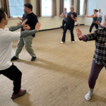 An image from a Tai Chi Class at Gorton Center. Instructor Ken Ning is up at the front of the classroom posed in a lunge with his arms straight out in front of him. Facing him are 10 students holding the same position
