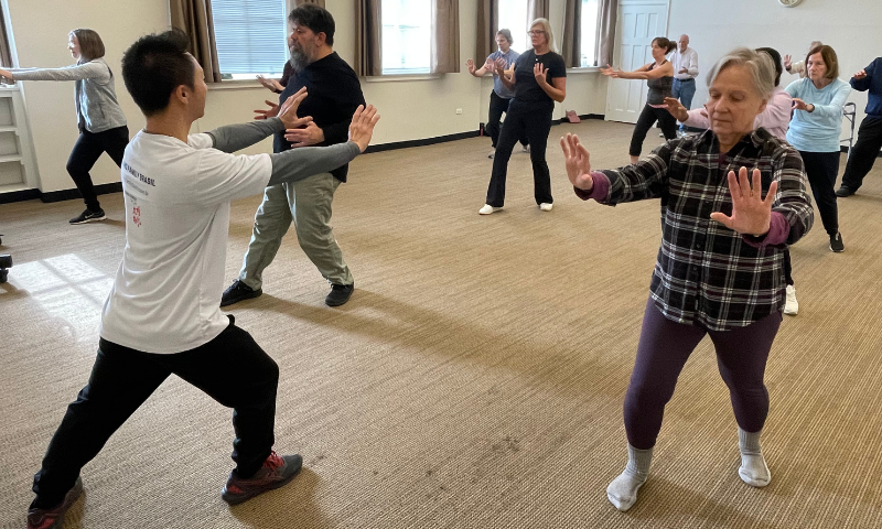 An image from a Tai Chi Class at Gorton Center. Instructor Ken Ning is up at the front of the classroom posed in a lunge with his arms straight out in front of him. Facing him are 10 students holding the same position