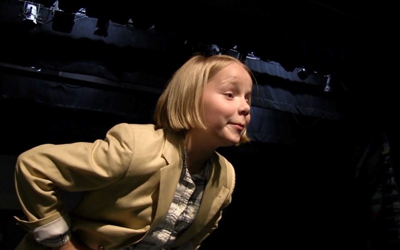 A young girl on stage leaning forward, singing to the audience