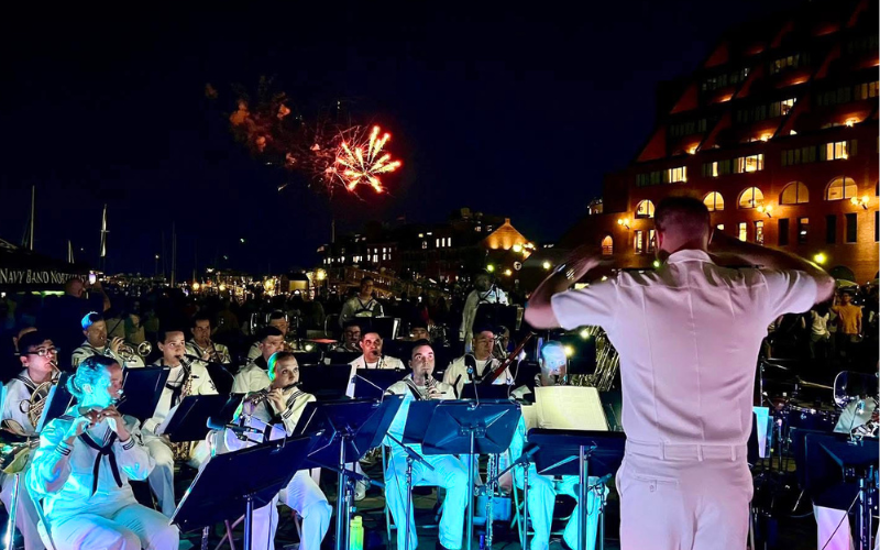 An image of the Navy Band Great Lakes performing at an outdoor concert. They are dressed in their white sailor uniforms. There is a large crowd around them and a firework is in the sky behind them