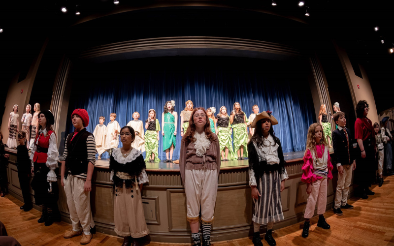 A large group of kids standing on and in front of a stage with a blue curtain drawn behind them. About half of the group are dressed as pirates and the other half as mermaids. They are all standing faced forward, about to take their bows. The image is show in panoramic mode