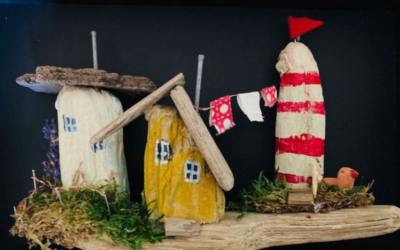 A set of 3 hand crafted, miniature wooden structures sitting on a piece of a wedgewood. There is moss by the base of the structures and a tiny bird sitting next to one. There are nails and small pieces of wood on top serving as the 'roof' of the mini houses.