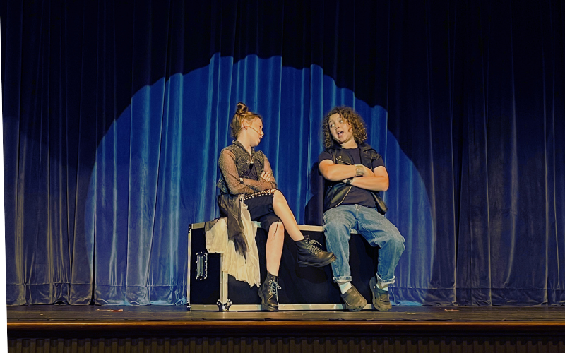 A boy and girl on stage sitting on top of an equipment box lit by a spotlight. They are dressed in rock n' roll style clothes and are looking at each other as they sing