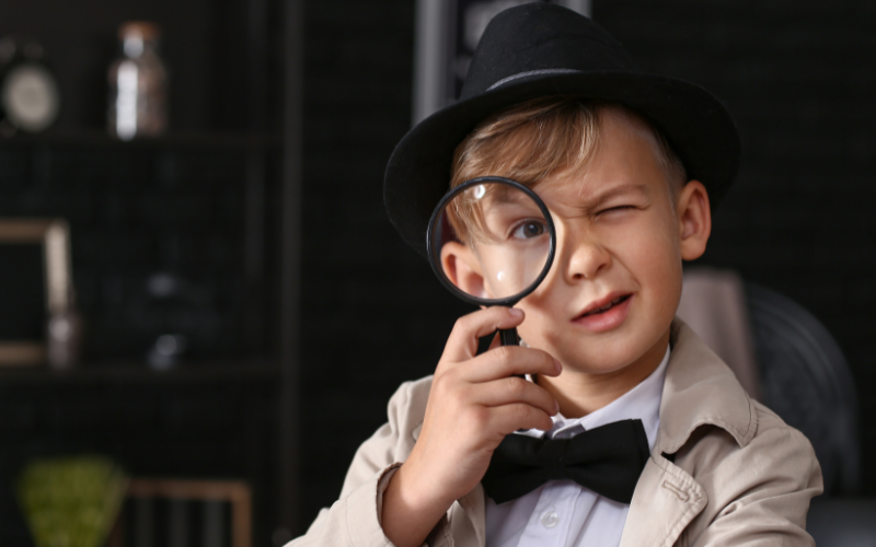 An image of a young boy dressed as an old timey detective. He is wearing a trenchcoat, fedora, bowtie and is looking through a magnifier glass.