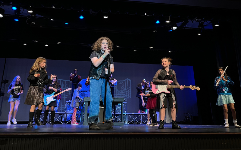 An image of a group of 12 kids performing on stage. They are all dressed in rock n' roll style with leather vests and jackets, torn jeans, sequins, etc. They are each holding an instrument except for the boy in the very front who is singing into a microphone.