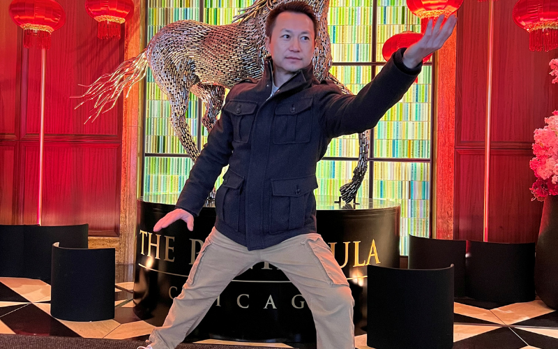 An image of Tai Chi instructor Ken Ning. He is posing with one arm up in front of his face and the other down to his side in front of the horse sculpture at the Peninsula Hotel in Chicago.