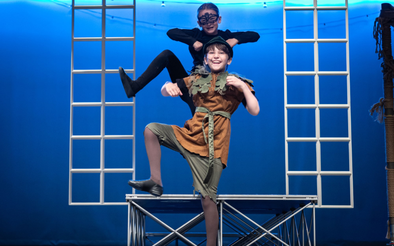 An image of two kids performing onstage. A boy is dressed as peter pan and is dancing with his arms up. Behind him is a girl dressed in all black with a mask over her eyes. She is doing the same dance move as the boy.