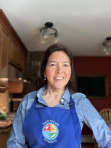 An image of Thais Boddick, wellness educator with Food for Life. She is standing in a kitchen wearing a blue apron with the Food for Life logo.