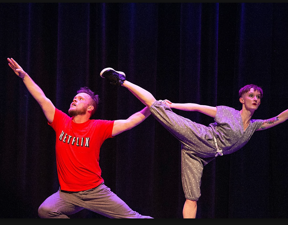 An image of comedy dance performers on stage in Happy Dance. The image has two performers; one is stretched in a lunge position and holding the leg of the person next to them who is in an 'arabesque' position. They both have serious expressions on their faces.