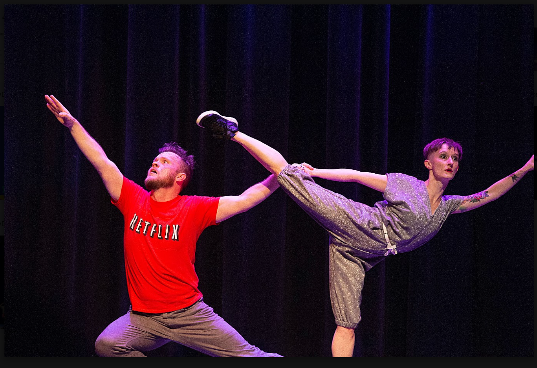 An image of comedy dance performers on stage in Happy Dance. The image has two performers; one is stretched in a lunge position and holding the leg of the person next to them who is in an 'arabesque' position. They both have serious expressions on their faces.