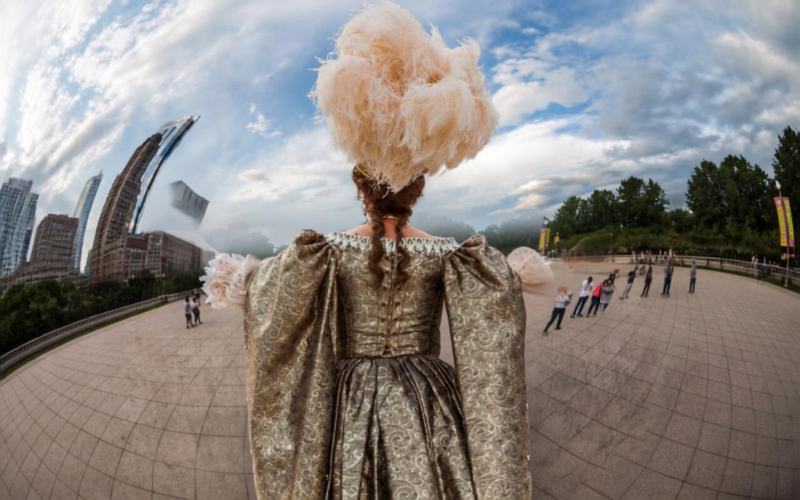 A wide angle image of a woman standing outside in a plaza. Her back is to the camera and she is wearing a large, ornate dress with a large feather piece in her hair. Her arms are outstretched.