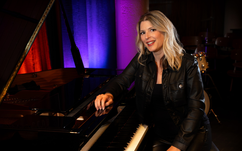 A picture of Jo Ann Daugherty sitting at a grand piano. She is wearing all black, smiling and looking at the camera. Behind her, multi colored lights shine against curtains.