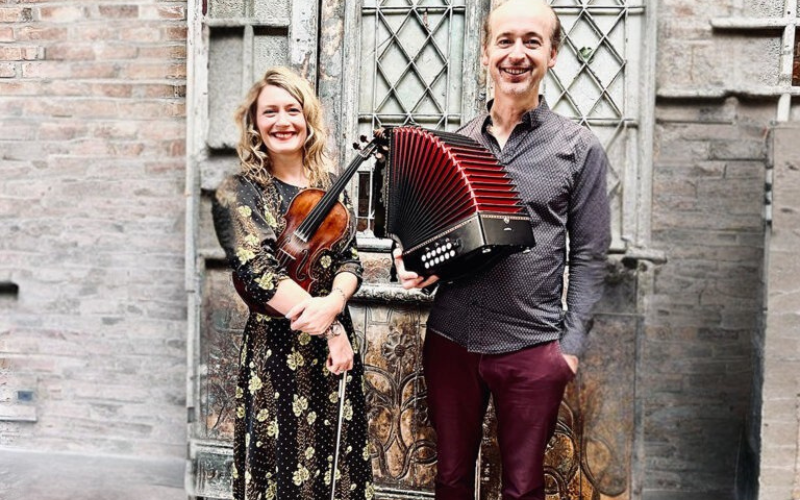 Katie Grennan and John Williams are standing outside of a brick building standing holding their instruments. They are smiling and looking directly at the Camera