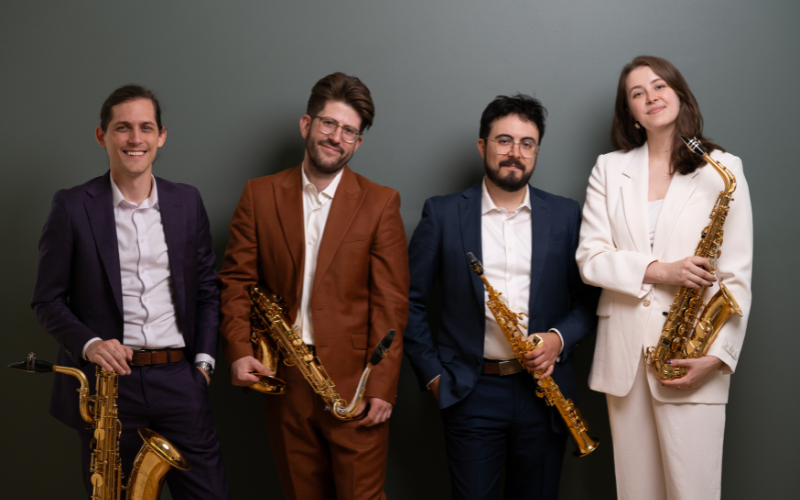 A portrait of the 4 members of the Nois Saxophone Quartet. They are all well dressed in suits, smiling and looking at the camera while holding their saxophones