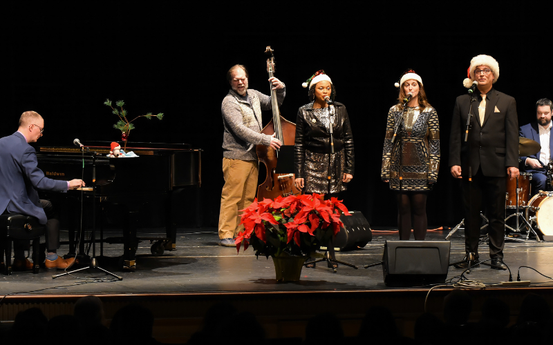 An image of the Chris White Trio performing Peanuts Jazz on stage at Gorton Center. There is a large poinsettia at the front of the stage in the center and the band and singers are staggard around the stage- some of them are wearing Santa Hats.