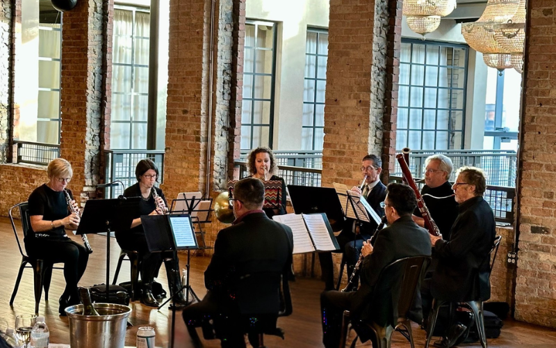 A photo of Strings attached performing. They are in a large, open room with brick walls and lots of windows. They are all wearing black and seated in a semicircle.