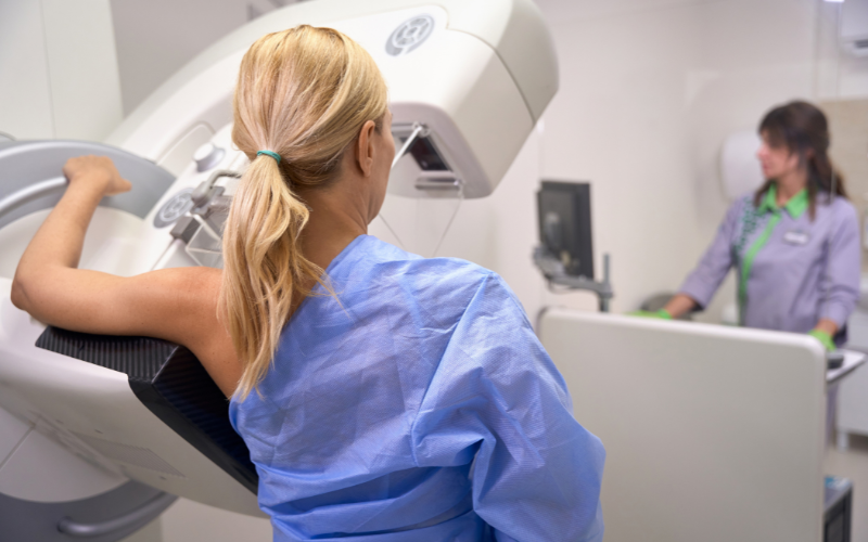 Blonde woman stands at mammography unit while technician runs the test during her annual exam for Breast Health