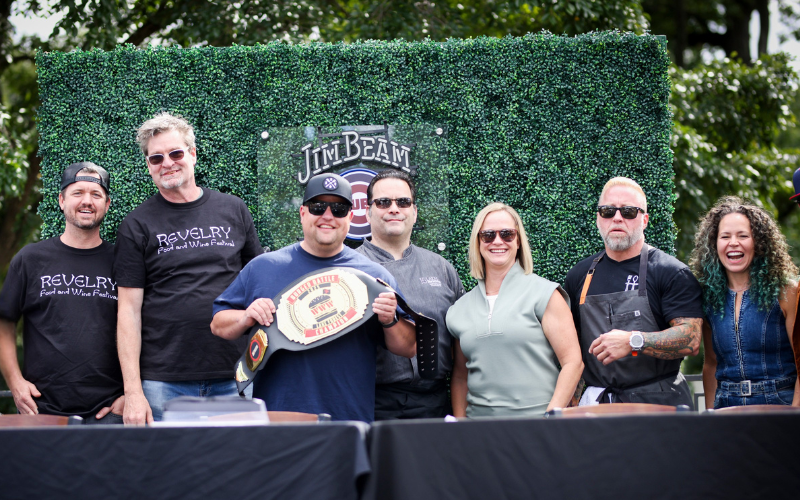 The Judges, host and winners of the Burger Battle are posed together standing behind the judges table. They are smiling and looking at the camera and the winner is proudly holding the championship belt