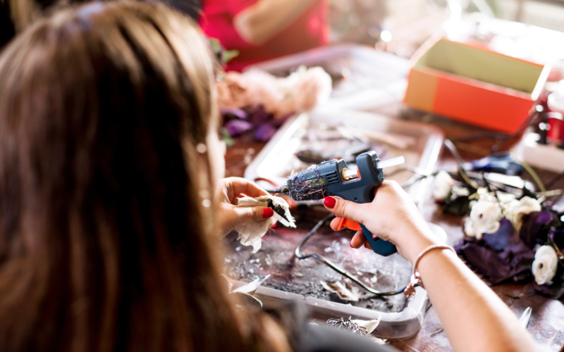 An image of a woman using a glue gun during Community Craft Night. The camera is behind her and on the table in front of her are fake flowers and other crafting materials