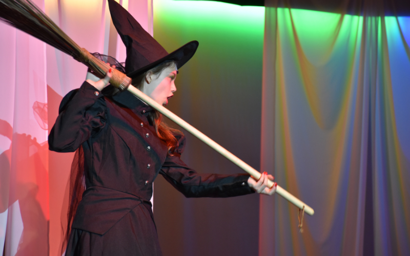 An image of a young girl on stage dressed as the wicked witch of the west from 'the wizard of oz'. She is holding her broom up as if to use it as a weapon. Behind her, the back of the stage is light with green and pink lights