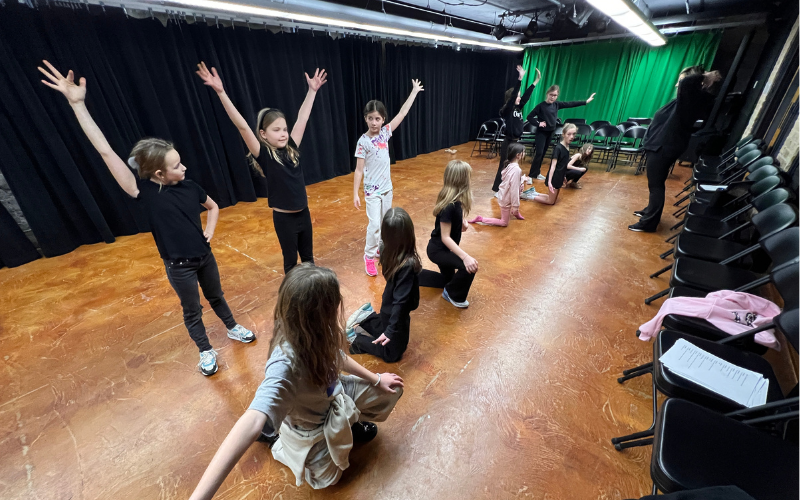 A group of children are in a black box theater doing choreography in explore theater basics class. Their director is giving them instruction